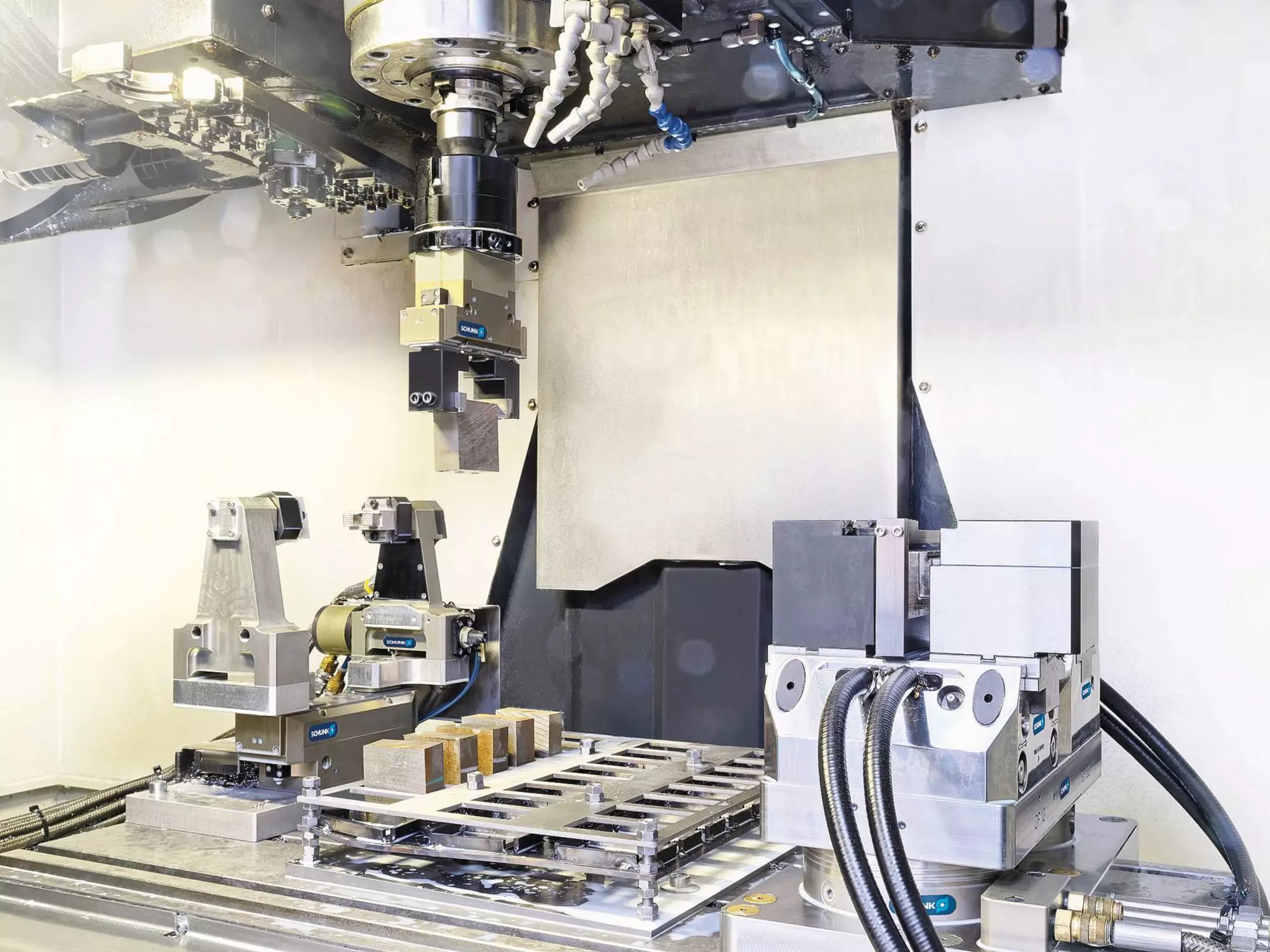 Inside a machining center: a pneumatic clamping station with its supply hoses, a raw-part tray and a gripper held in the spindle